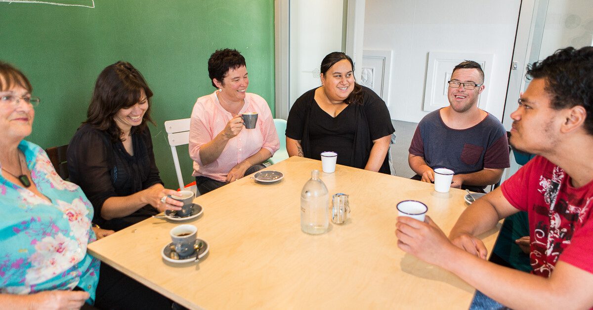 Group of six people sit around a table drinking coffee and talking