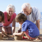 Two grandparents kneeling on the beach making sandcastles with two children.