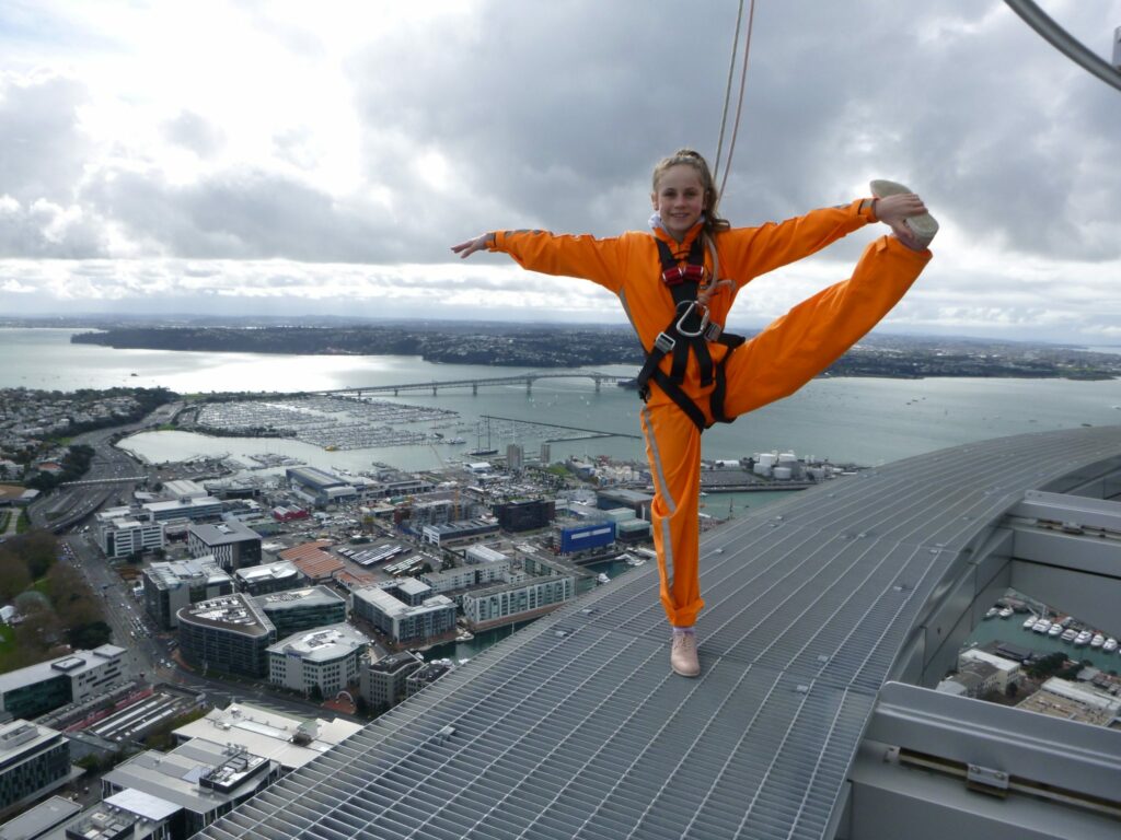 Young woman in orange suit does gymnastic poses on the edge of the Auckland Sky Tower.