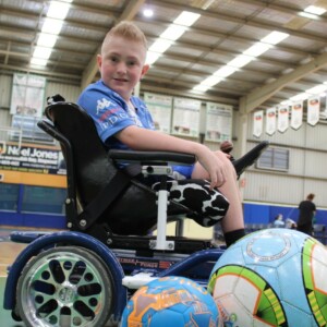 Twelve-year-old Angus, with short blonde hair, sits in hit football powerchair while looking at the camera. He is in a school gymnasium. He has footballs around him.