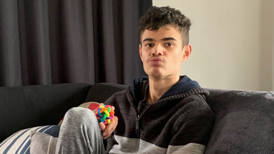Tyler has short, dark curly hair and is sitting on the couch at home. He is wearing a jersey and trackpants and holding a colourful fidget ball.  