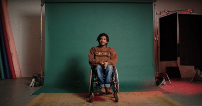 Shakti wears a chocolate brown sweater and blue jeans, and sits in a wheelchair.