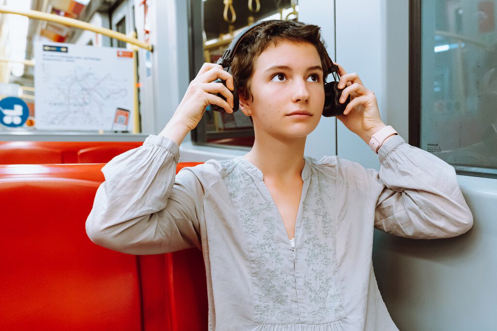 A teenage girl with short cropped hair wearing wireless headphones, is riding train or subway car, enjoying music.