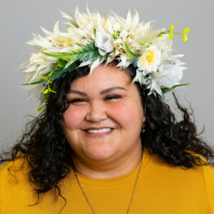 Te Aroha is wearing a yellow shirt and a flower crown. She is smiling brightly.