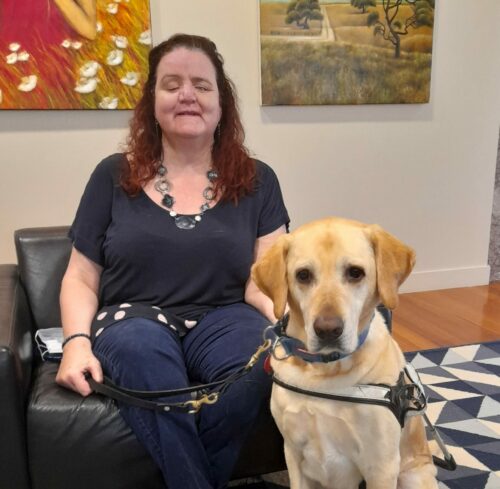 Martine Abel-Williamson sits on a chair next to her guide dog, Greg. Martine wears a black shirt and a chunk necklace. She has red hair.  Greg is an alert looking golden retriever.
