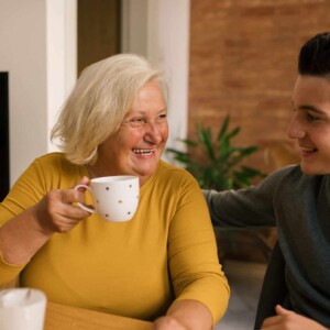 A woman in a mustard shirt is drinking coffee. She is smiling at her grandson, who is sitting beside her.